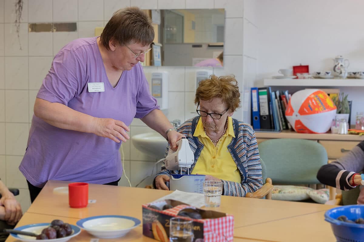 Pflegerin hilft einer Seniorin beim Teig rühren